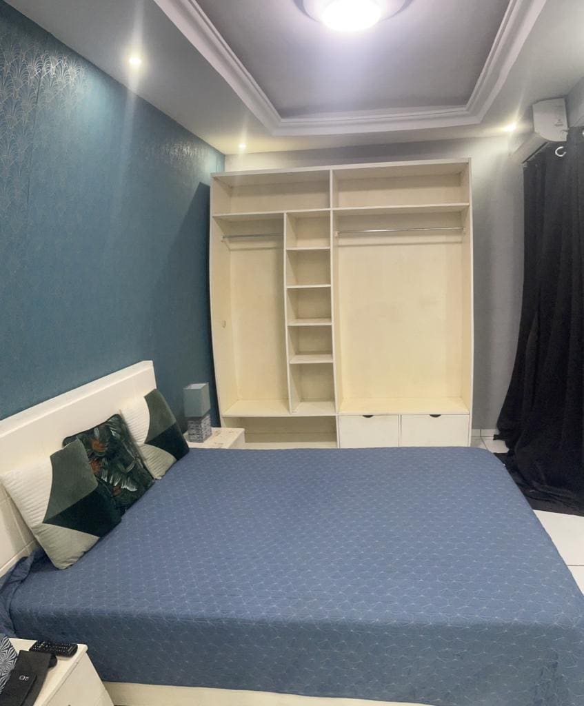 The sleeping area features a spacious bed with a blue quilt. A light-colored wardrobe with open shelving is situated against a teal wall, while a dark curtain allows for light control. A bedside table is positioned beside the bed, contributing to a functional design.