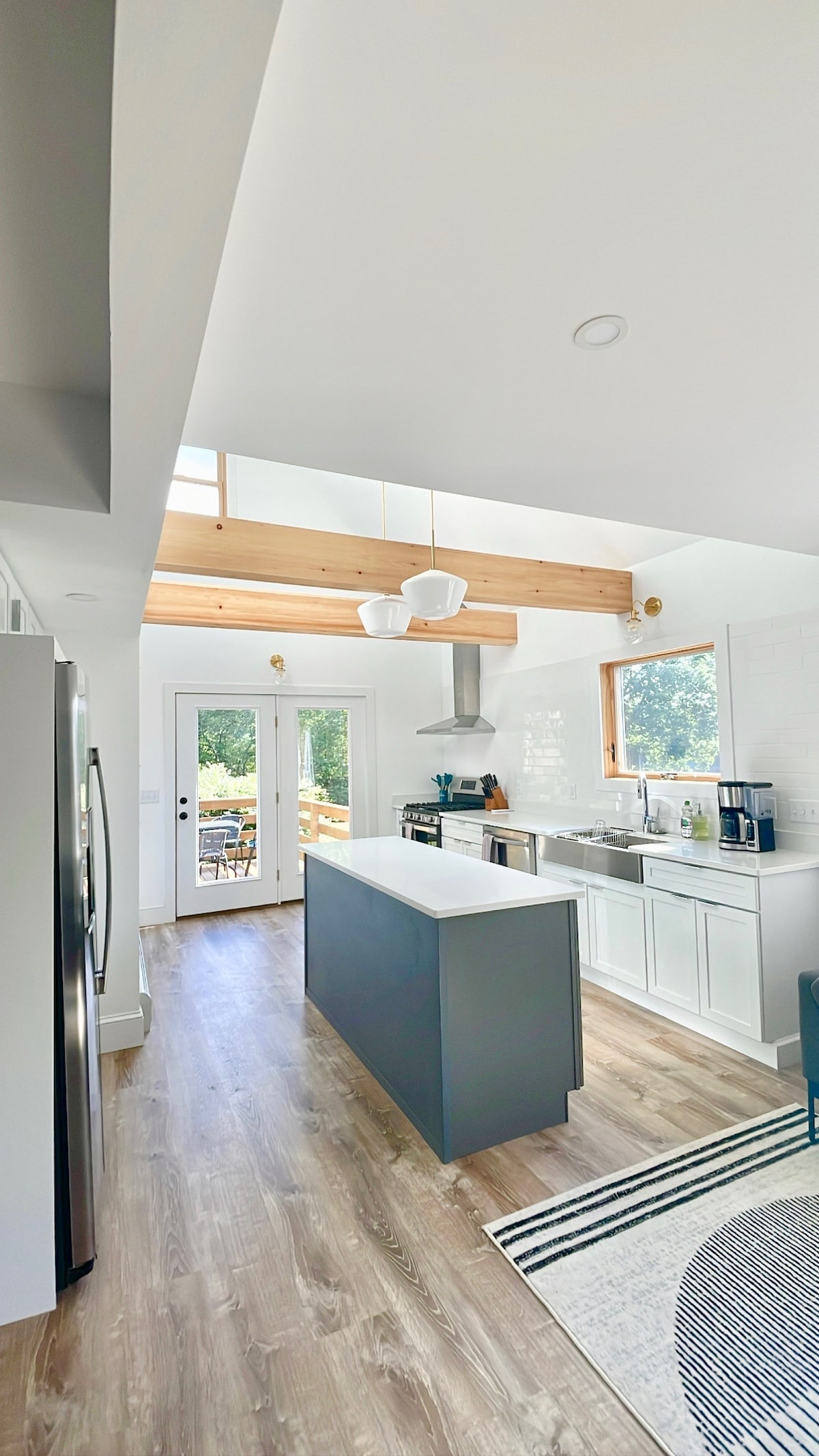 A modern kitchen is showcased with an island bar, complemented by sleek cabinetry and bright countertops. Natural light floods the space through large windows, illuminating the stainless steel appliances. A sliding door offers direct access to an outdoor dining area.
