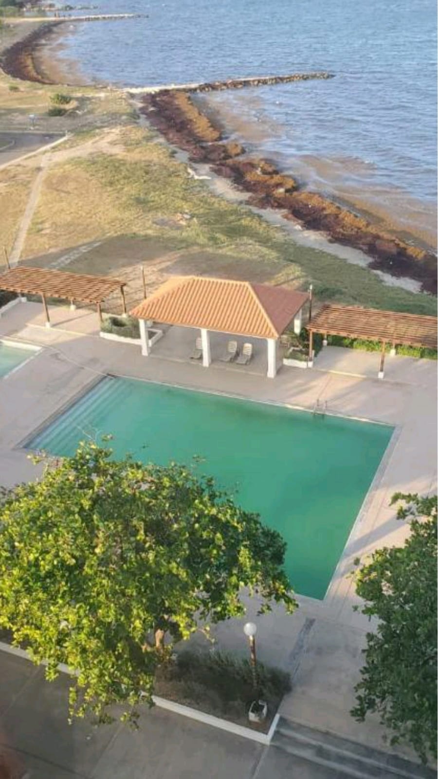 An outdoor pool is seen with clear turquoise water, bordered by a spacious deck. A gazebo with seating is positioned nearby. The coastline is visible in the background, featuring sandy areas and the gentle waves of the water.