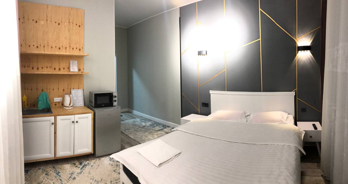 A well-lit guest room features a spacious bed with white linens. A kitchenette with a microwave and refrigerator is positioned against a wooden wall. The decor includes geometric lighting on a gray accent wall, adding a modern touch to the inviting space.
