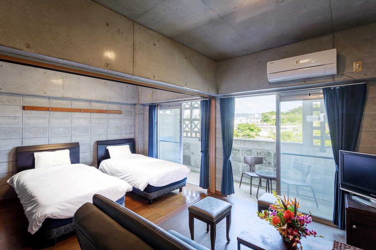 The spacious room features two single beds with white linens, positioned beside a glass sliding door that leads to a balcony. A small table and chairs are set by the window, with a vibrant arrangement of flowers on the floor. Air conditioning and a television are also present.