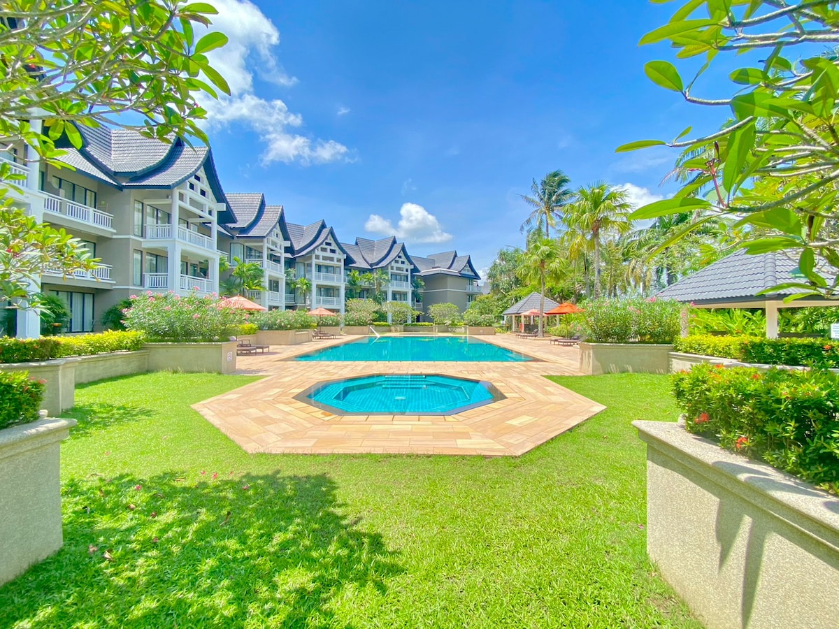 The outdoor area features a spacious swimming pool surrounded by lush greenery. A smaller kid’s pool is visible, complemented by manicured lawns and tropical plants. The pagoda-style buildings create a welcoming backdrop beneath a clear blue sky.