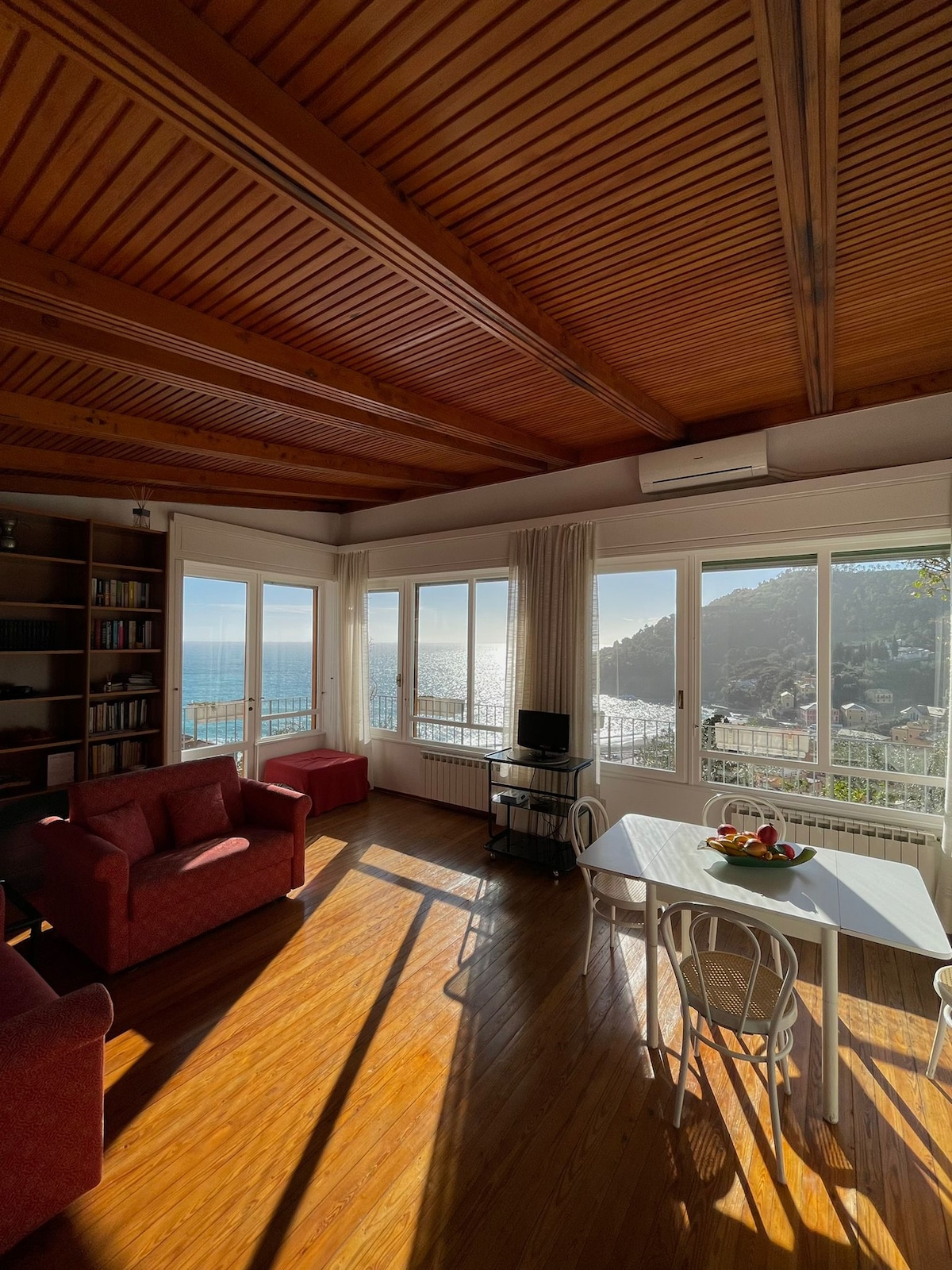 A bright living area features large windows with a panoramic view of the sea. Natural light floods the space, highlighting the wooden ceiling and warm-colored furnishings. A dining table sits adjacent to a comfortable seating area, creating an inviting atmosphere for relaxation.