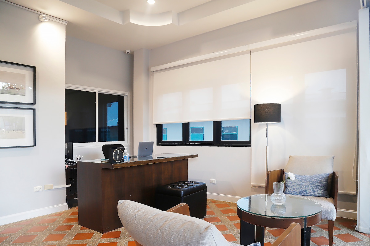 A modern workspace is showcased, featuring a wooden desk paired with a laptop and clock. A comfortable armchair and a glass-topped table are positioned nearby, while large windows provide natural light, complemented by soft white blinds.