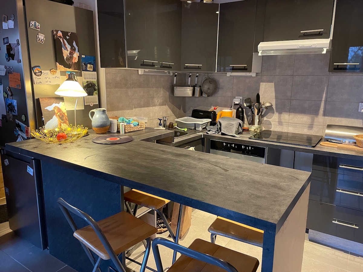 The kitchen features modern cabinetry in dark tones, equipped with stainless steel appliances. A large countertop provides space for meal preparation, while a bar area with two wooden stools offers a casual dining option. Soft lighting illuminates the workspace and decorative accents.