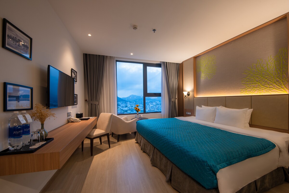 The room features a spacious bed with a turquoise coverlet, complemented by soft white pillows. A large window offers a view of the city and mountains. A seating area is present beside a wooden desk, and a wall-mounted television is positioned across from the bed.