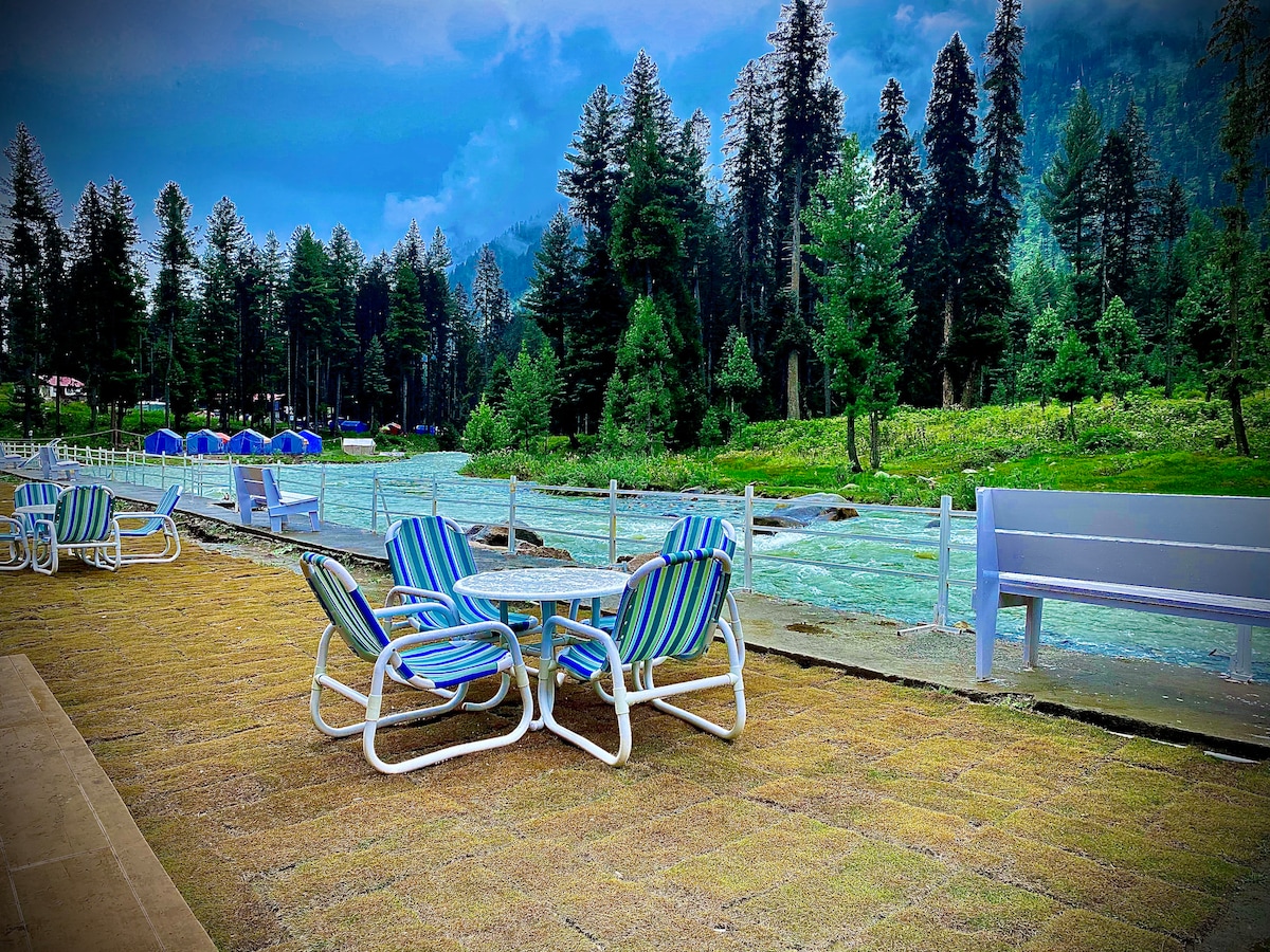 A riverside area features several lounge chairs and tables arranged on a grassy surface. Lush trees rise in the background, alongside the flowing river, creating a serene outdoor setting.