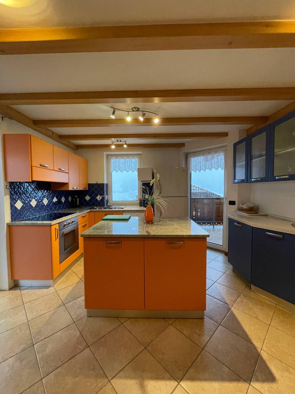 A spacious kitchen is presented with a central island and contrasting orange and blue cabinetry. Natural light filters through two windows adorned with lace curtains, illuminating the tiled floor and stone countertops. Stainless steel appliances are integrated, providing a modern cooking space.