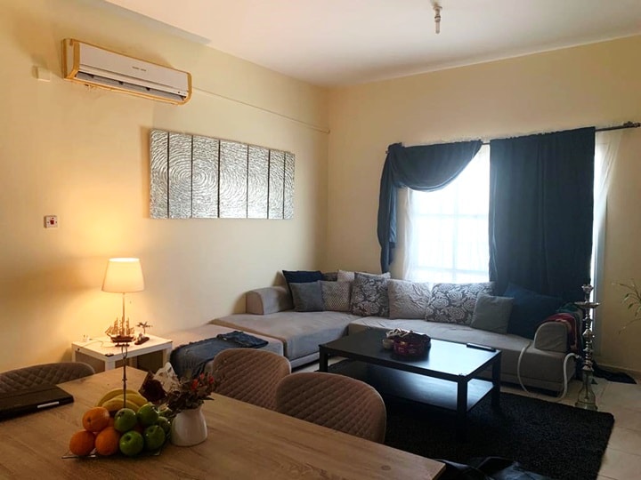 A bright living area features a spacious sectional sofa, complemented by a wooden coffee table. A decorative piece hangs above the sofa, and light flows through the window adorned with dark curtains. A small dining table with fruit adds to the inviting atmosphere.
