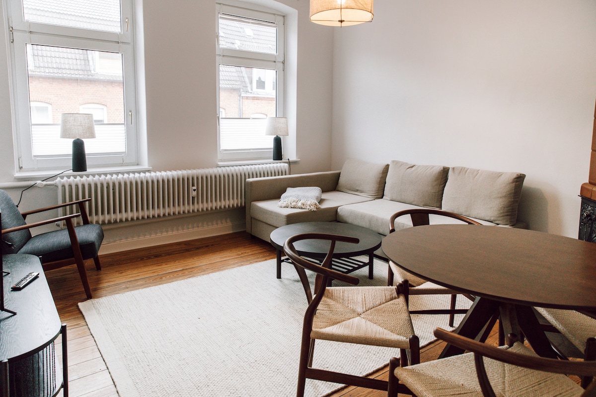 A spacious living area is highlighted by natural light from multiple windows. A comfortable sofa complements a round dining table surrounded by chairs. A cozy rug lies beneath the furniture, while the room features warm wooden flooring and clean, neutral-colored walls.