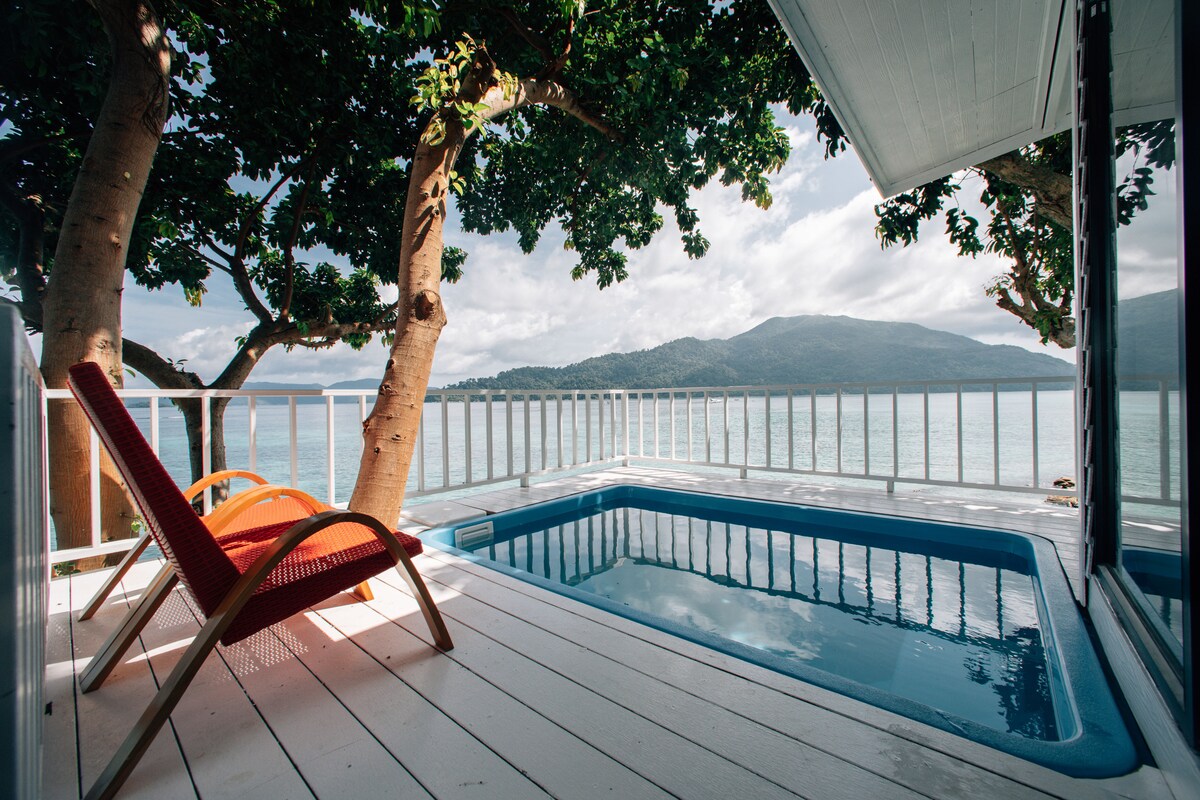 A sunlit outdoor area features a private pool framed by wooden decking. A bright orange lounge chair is positioned to enjoy views of the water and surrounding mountains, with lush green trees providing natural shade.