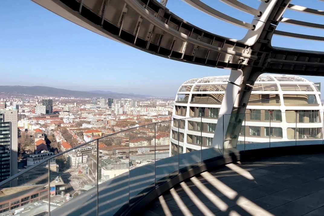 A panoramic view showcases a vibrant cityscape from an elevated balcony. The sleek structure with curved elements contrasts against the skyline, while sunlight casts dynamic shadows across the spacious terrace, highlighting the open and airy atmosphere.