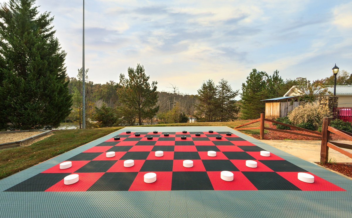 An outdoor giant checkerboard is displayed on a paved surface, featuring oversized black and white pieces. The surrounding area includes trees and a pathway leading to buildings in the background, contributing to a recreational atmosphere.