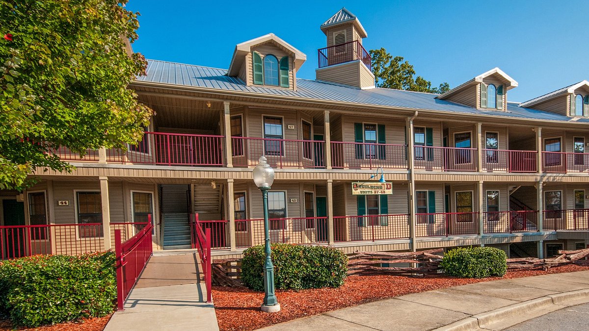The exterior of a multi-unit building is shown, featuring a light-colored façade with red accents. A sloped metal roof and a central tower are visible, along with red railings on the balconies. Surrounding greenery and a paved walkway enhance the inviting entrance.