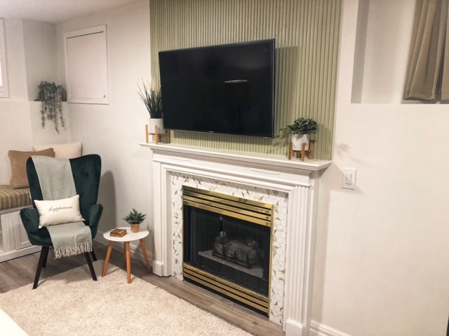 A living area features a modern fireplace with an ornate design beneath a large television. Indoor plants are placed on top of the mantel and beside a cozy green chair. Natural light enters through nearby windows, enhancing the relaxing atmosphere of the space.