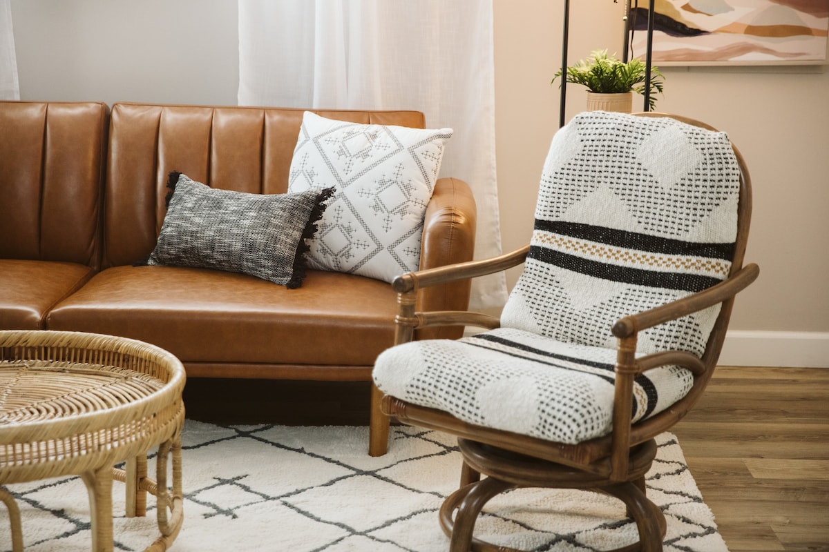 A cozy seating area features a brown leather sofa accompanied by a patterned pillow. A woven armchair with a cushion and a round rattan coffee table sit atop a light area rug. Natural light enters through the nearby window adorned with sheer curtains.