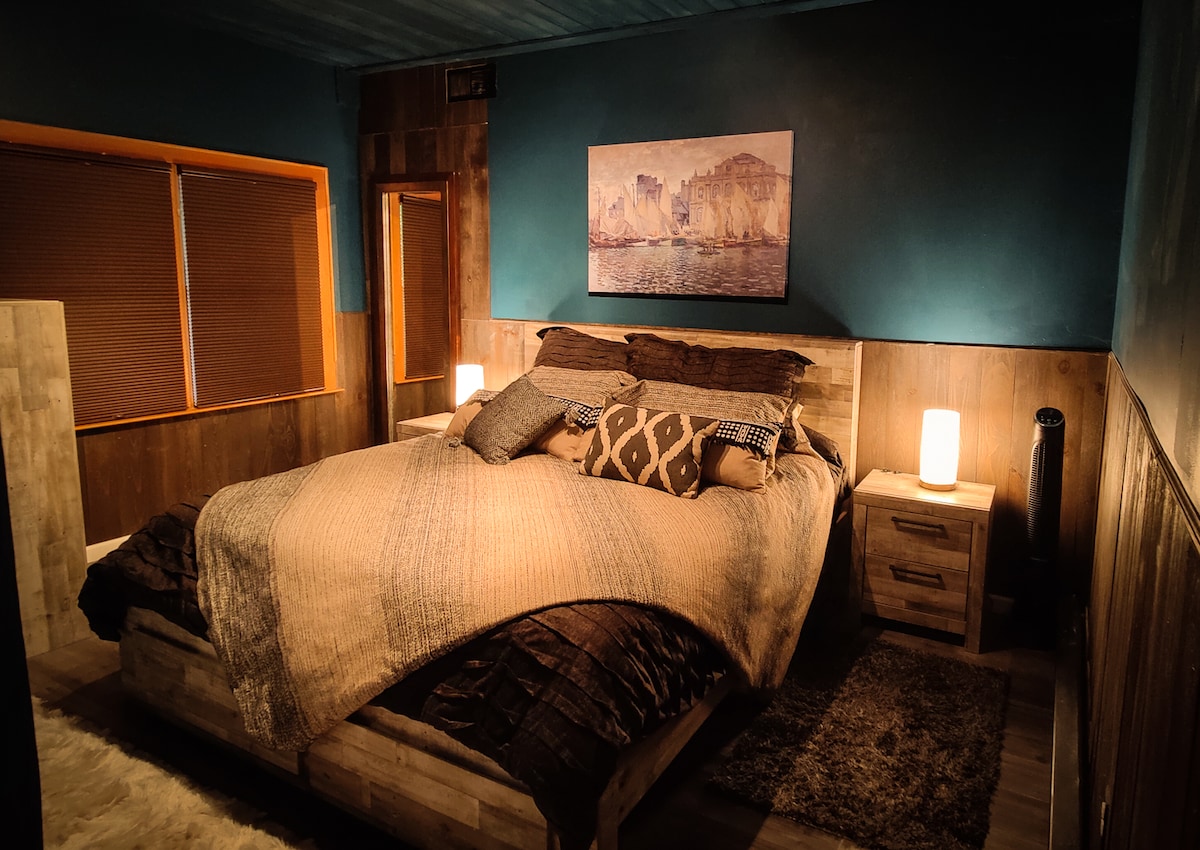 A tranquil bedroom features a spacious bed covered in soft, layered linens and decorative pillows. Warm lighting is provided by two bedside lamps, and artwork hangs above the bed. Large windows with wooden blinds allow for natural light, complementing the calming blue walls.