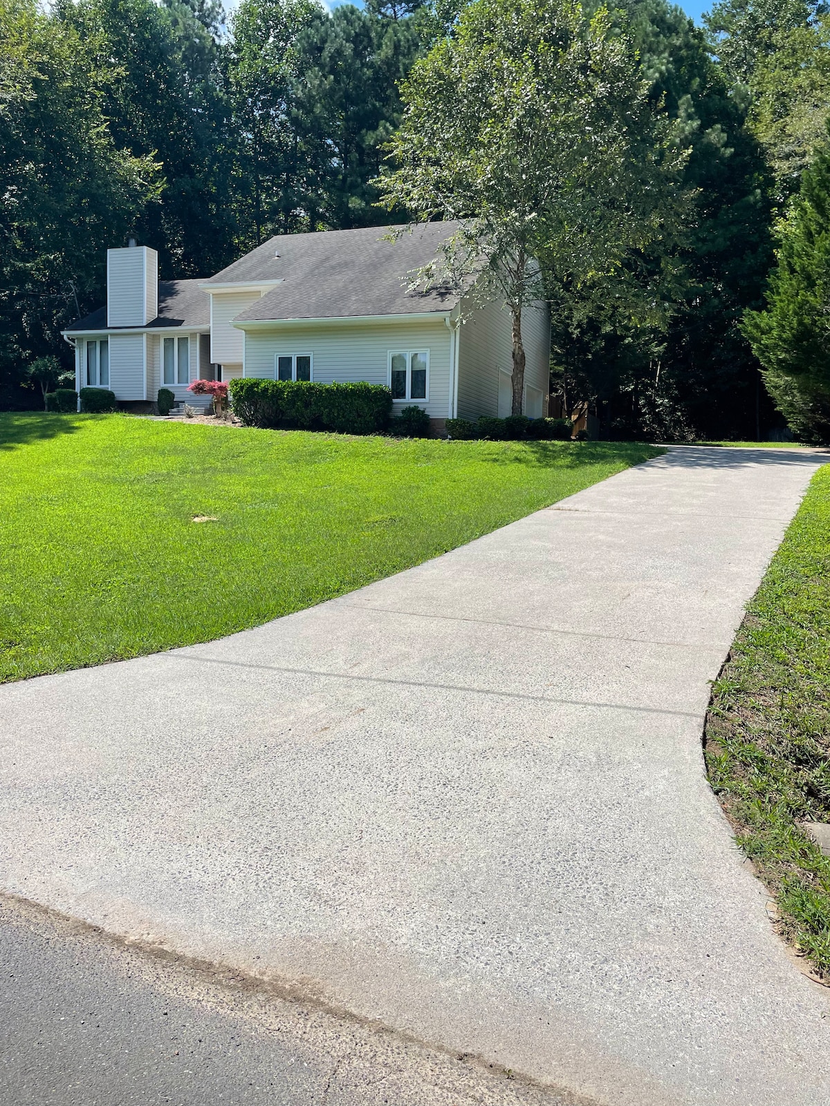 A well-maintained driveway leads to a two-story house, surrounded by lush greenery and trees. The inviting front yard features a neatly trimmed lawn and colorful flower beds, enhancing the overall peaceful ambiance of the property.