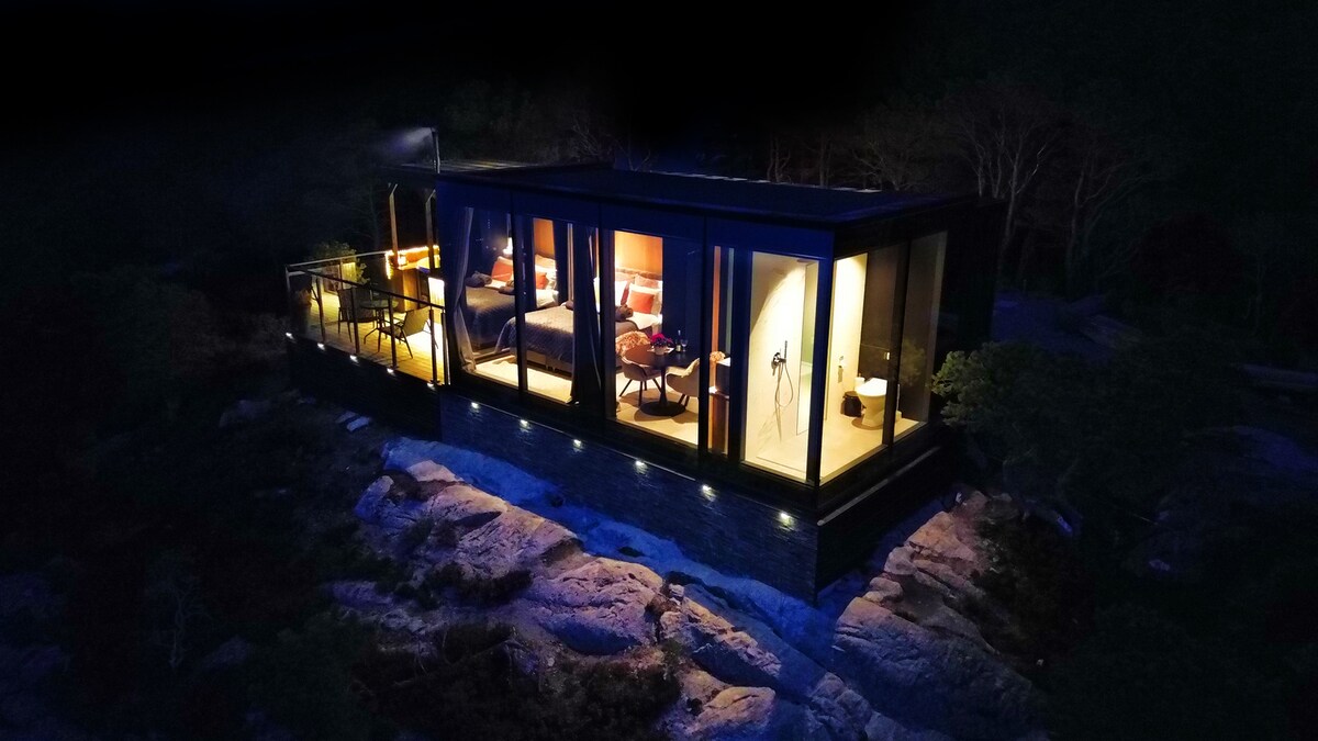 A modern cabin is illuminated at night, featuring large glass walls showcasing a cozy sleeping area. The structure is surrounded by natural rocky terrain, with outdoor seating visible on the balcony. Soft lighting accentuates the cabin's design against the dark sky.