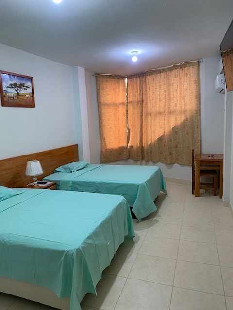 A bright guest room is presented with two spacious beds covered in light blue linens. A bedside table with a lamp is positioned next to each bed. Natural light enters through a large window adorned with sheer, patterned curtains, creating a welcoming atmosphere.