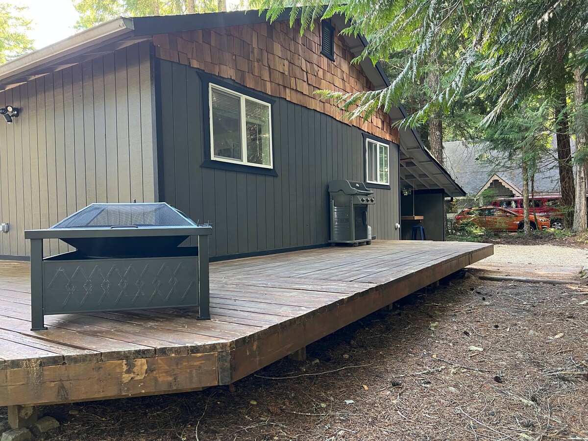 The cabin exterior features a spacious wooden deck surrounded by tall trees. A black fire pit is positioned on the deck, alongside a grill. Windows provide ample natural light, and the entryway is easily accessible from the outdoor space.