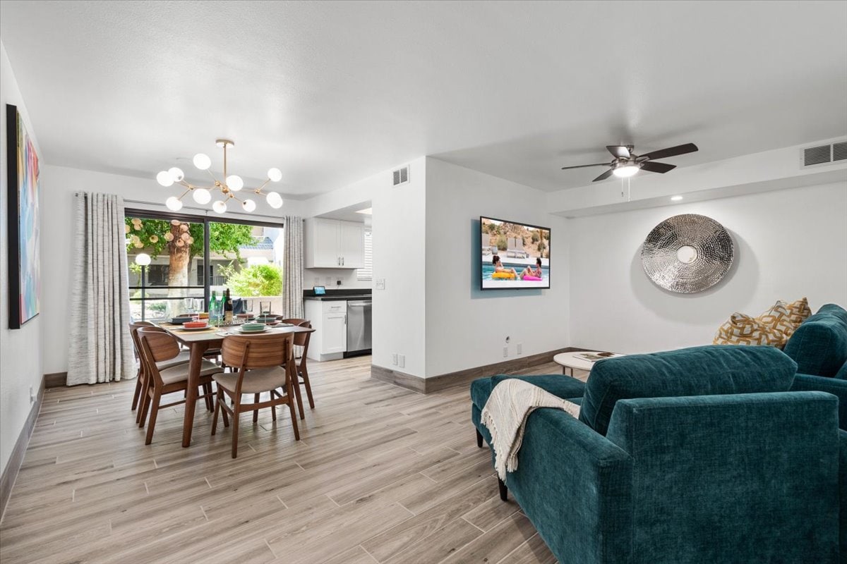 A spacious living area is shown, featuring a plush teal sofa and a light wood dining table surrounded by four chairs. A large wall-mounted television displays a vibrant image. Natural light filters through a window, highlighting the modern decor and open layout.