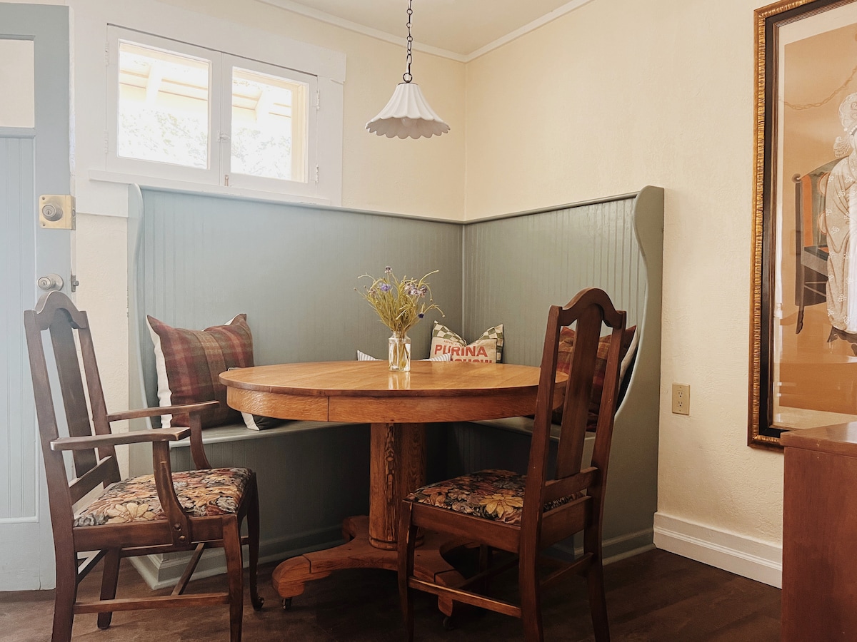 A dining area is highlighted by a round wooden table seated by four chairs. The wall features a soothing green panel, and natural light streams in through a window. A pendant lamp hangs overhead, providing a warm, inviting glow to the space.