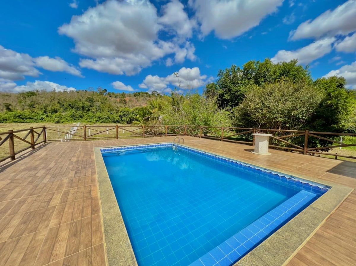 A clear blue pool is set on a wooden deck, surrounded by lush greenery and distant hills. The sky is bright with fluffy clouds, enhancing the outdoor space ideal for relaxation and gatherings.