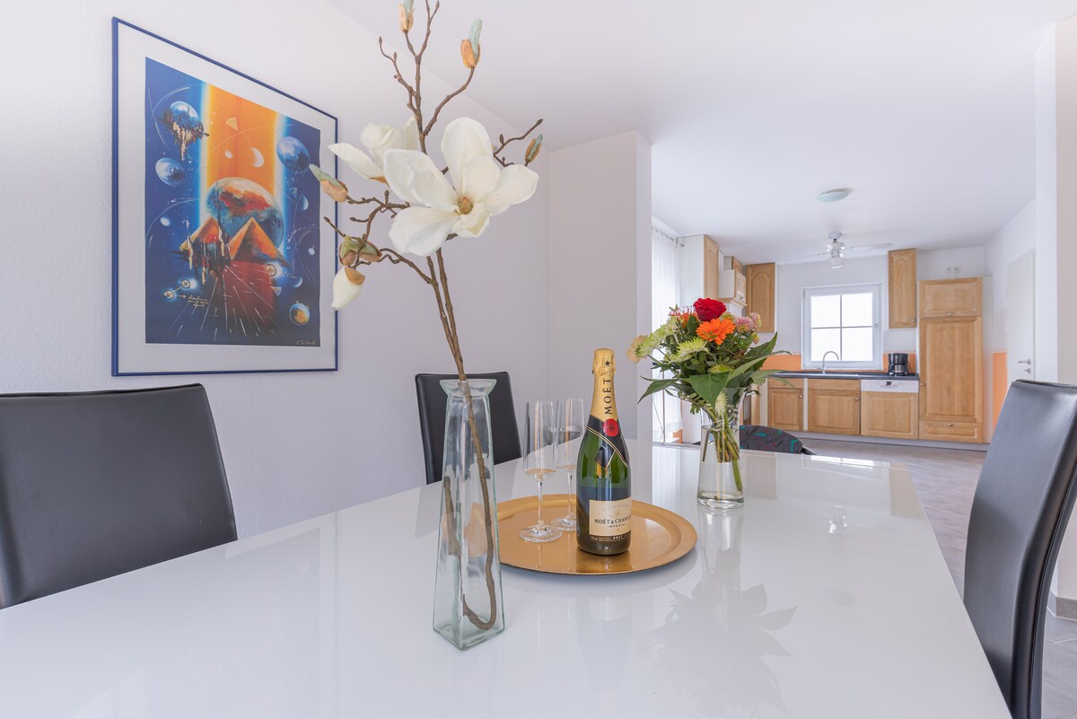 A dining table is set with two flutes and a bottle of champagne, accompanied by a vase of fresh flowers. An artwork depicting a space theme hangs on the wall. The kitchen can be seen in the background, featuring wooden cabinetry and natural light.