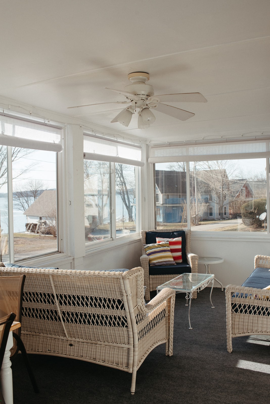 A sunlit enclosed porch is furnished with wicker seating and colorful cushions, creating a comfortable space for relaxation. Large windows provide views of the surroundings, while a ceiling fan is present for added comfort. A small glass table complements the seating arrangement.