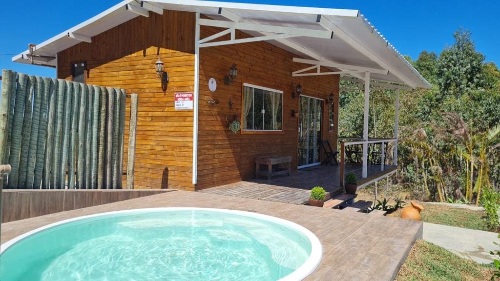 A modern wooden house with large glass windows is presented, surrounded by lush greenery. A circular pool is situated in the foreground, with a wooden deck leading to the entrance. The roof is designed with overhangs for shade, enhancing the outdoor space.
