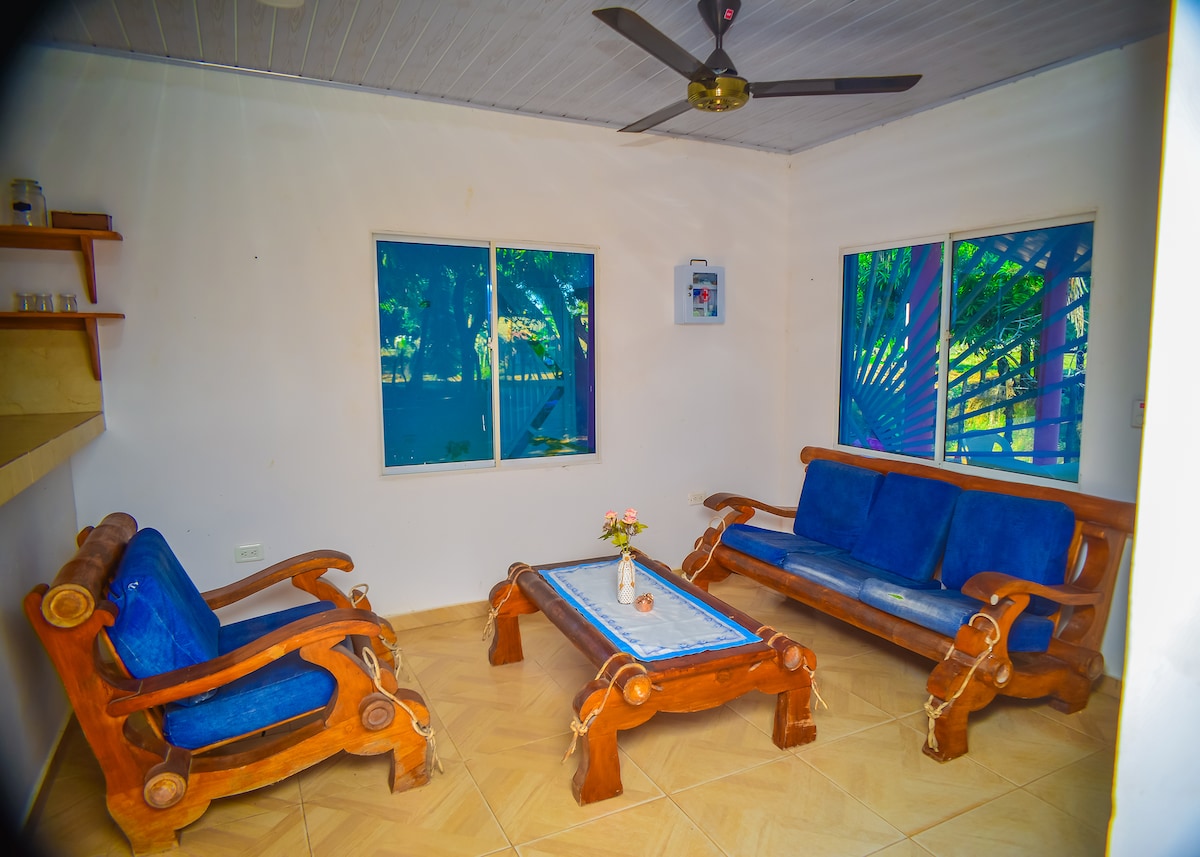 A living area features wooden seating with blue upholstery, arranged around a rectangular coffee table. Natural light enters through two large windows, which provide a view of the outdoors. A small decorative item sits on the table, adding a subtle touch to the space.