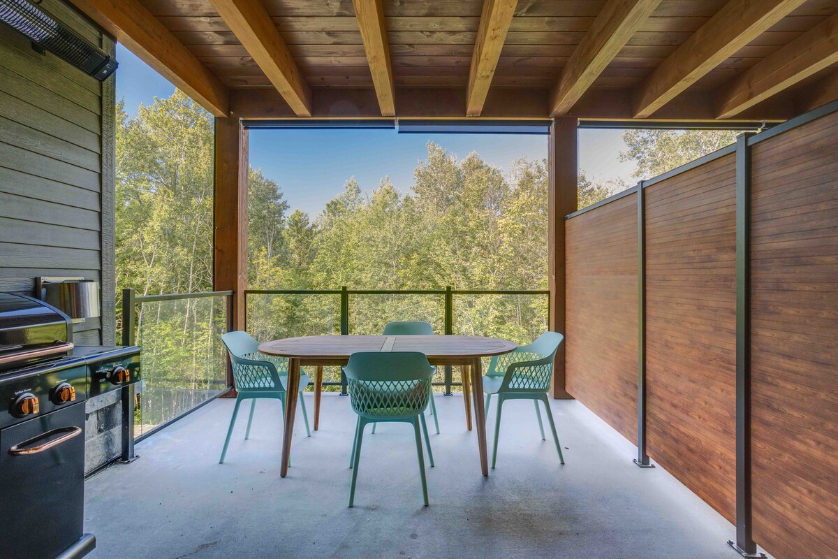A spacious terrace is showcased with a dining table and four chairs, surrounded by glass and wooden paneling. A barbecue grill stands nearby, with lush greenery visible beyond the railing, offering a tranquil outdoor experience.