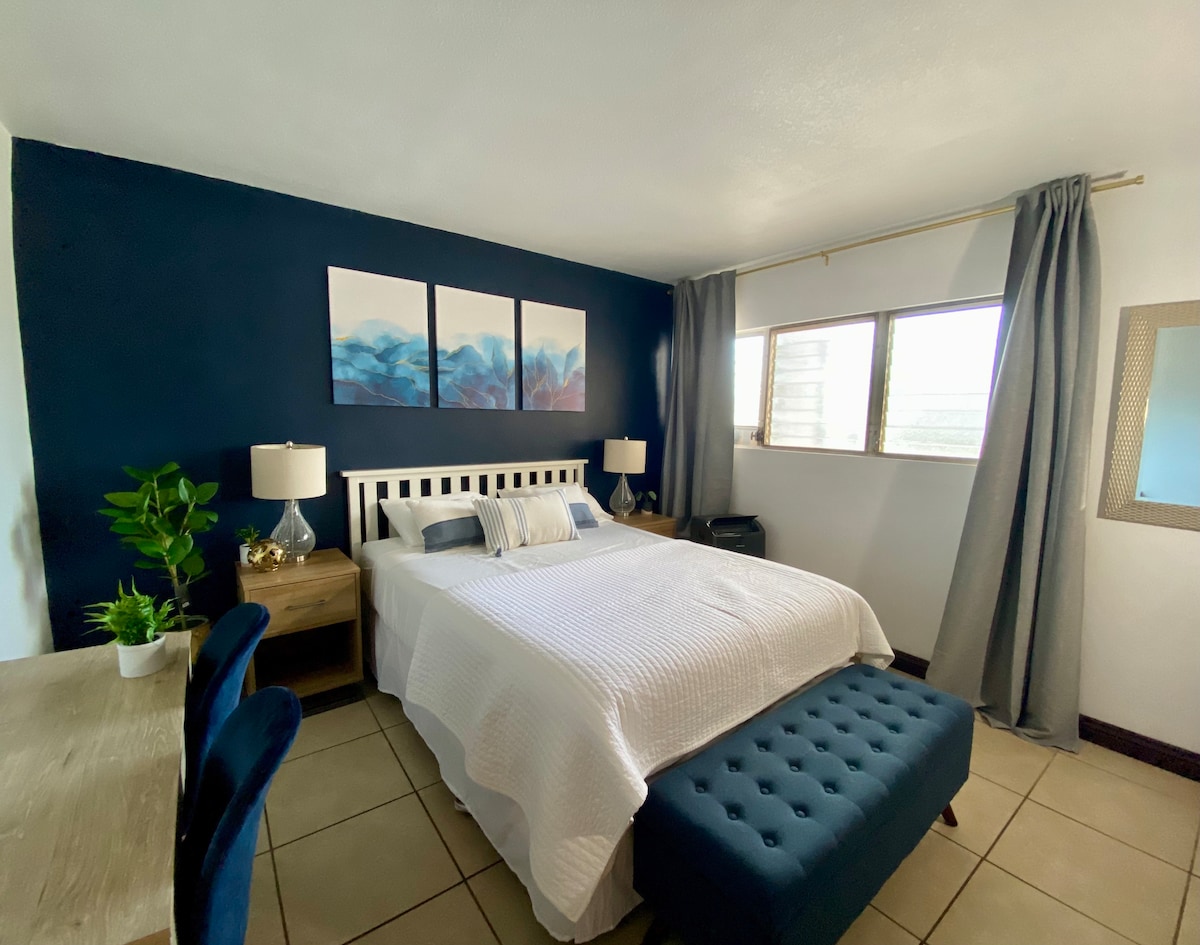 A bright studio space features a queen bed with a textured white coverlet, complemented by blue accent pillows. Artwork with mountain themes adorns the wall above the bed. Two lamps provide soft lighting, and a workstation with a plant and seating are positioned nearby.