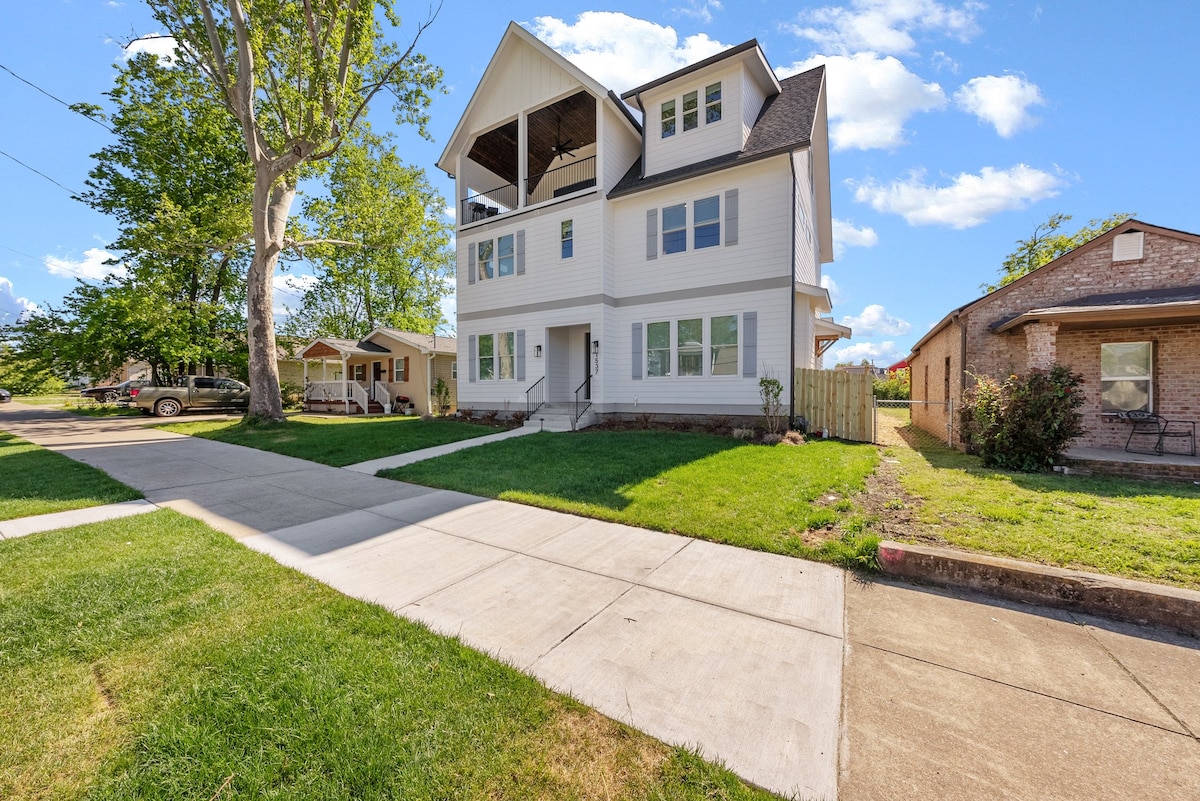 A three-story white house with a gabled roof is shown, featuring a large outdoor deck on the top floor. The front yard is well-maintained, with green grass and a paved walkway leading up to the entrance. Several trees and neighbors' homes are visible in the background.