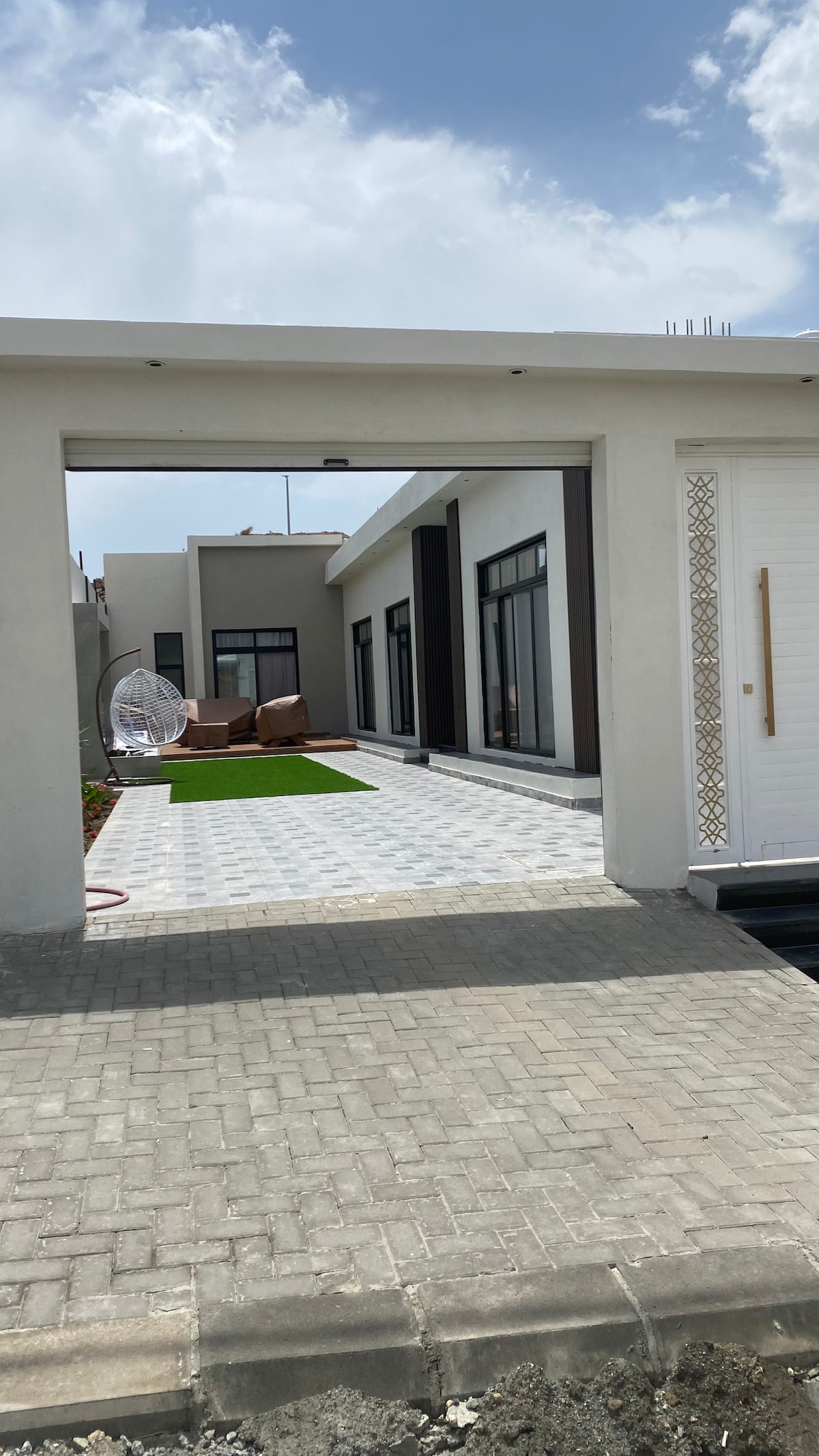 The exterior entrance opens to a paved pathway leading towards a modern home. A spacious outdoor area is visible, featuring lush green grass and seating arrangements. Large windows can be seen along the side of the building, allowing for natural light to enter the interior.