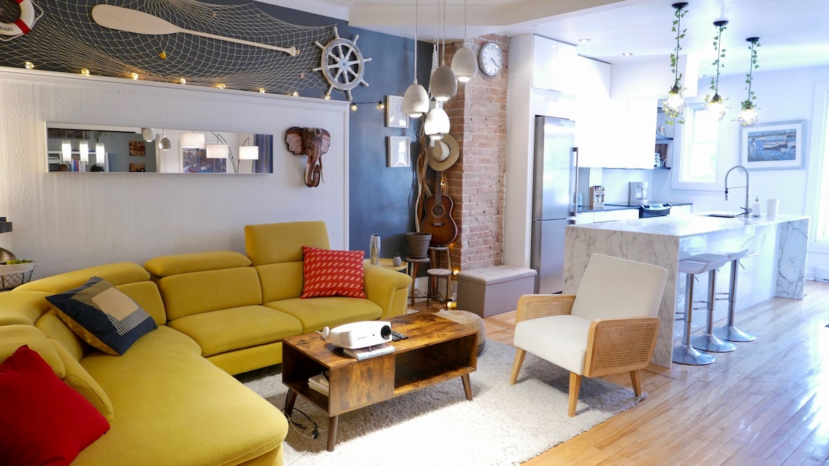 A spacious living area displays a large yellow sectional sofa with various cushions. A wooden coffee table is centered on a soft rug. A cinema projector is positioned near the wall. Modern kitchen elements, including a marble island and stainless-steel appliances, create a cohesive style.