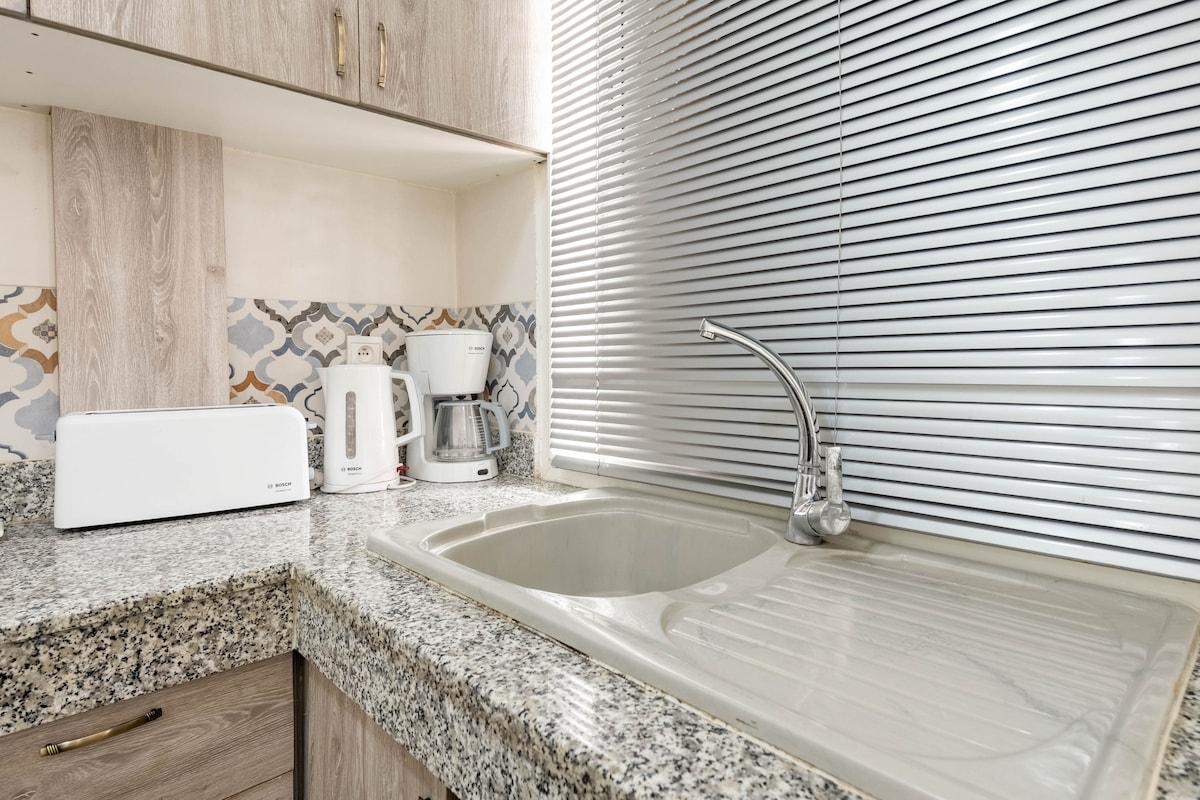 A well-equipped kitchen area features a spacious granite countertop with a sink. Small appliances, including a toaster, electric kettle, and coffee maker, are arranged neatly on the counter. Natural light filters through a window covered by horizontal blinds, enhancing the functionality of the space.
