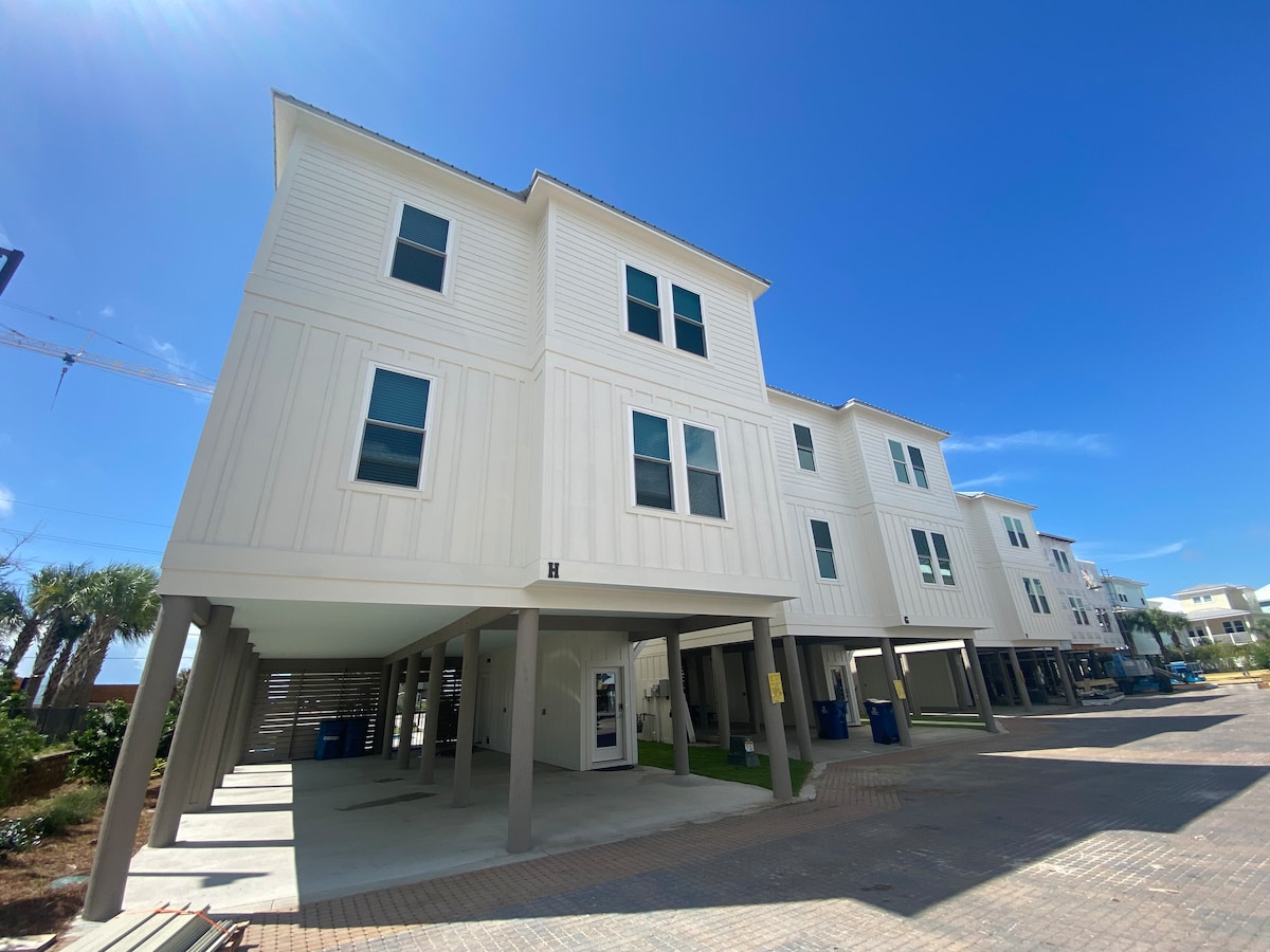 The three-story beach house features a modern design with white siding and large windows. The elevated structure rests on sturdy columns, creating an open parking area below. Lush palm trees and blue skies complement the welcoming atmosphere of the property.