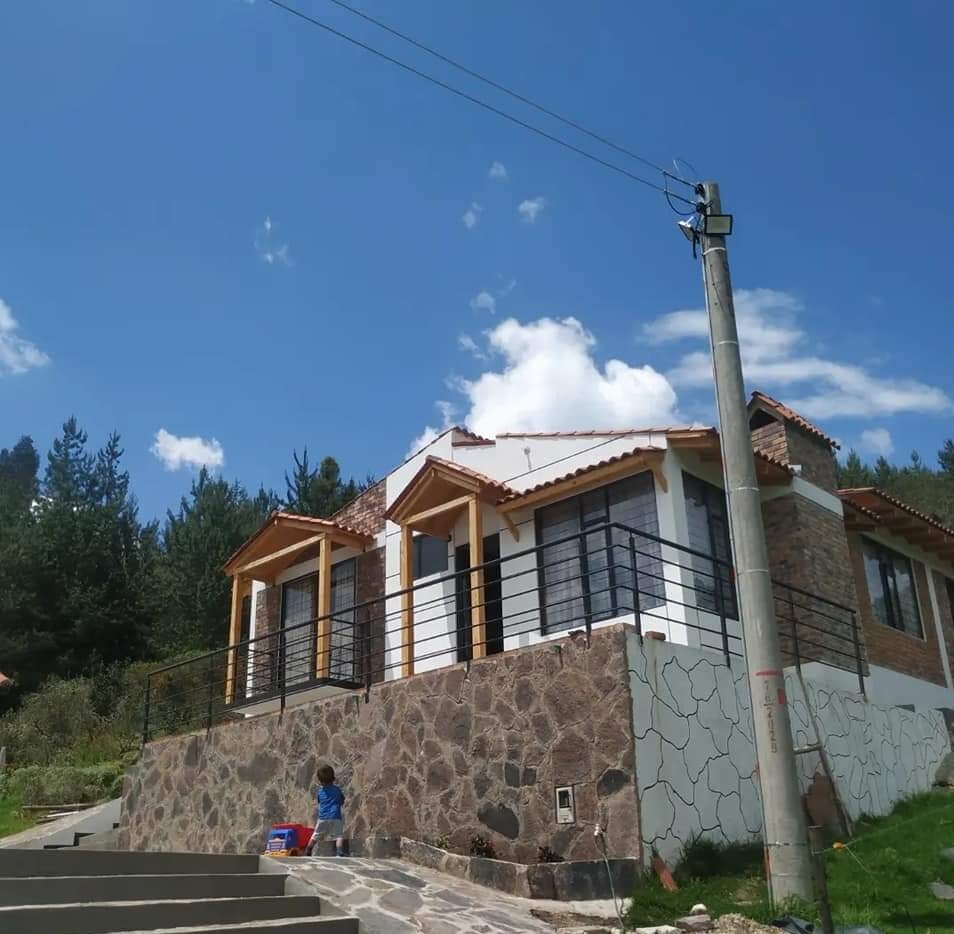 A single-story house features natural stone accents and a tiled roof. Two covered porches are supported by wooden beams, complemented by large windows. A child is visible playing on the steps, surrounded by greenery and a clear blue sky.
