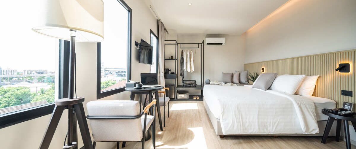 A well-appointed room features a spacious bed with light-colored linens, complemented by a minimalist design. A small seating area is arranged near a window, offering views of the surrounding area. A flat-screen TV and modern amenities are also visible, providing comfort and convenience.