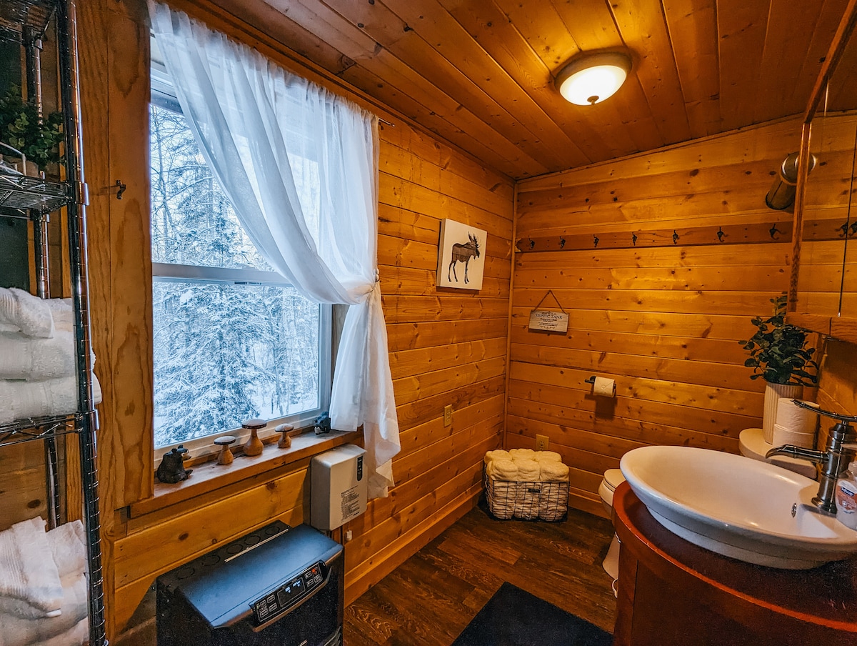 The bathroom features wooden walls and a large window with sheer curtains, allowing natural light to illuminate the space. A modern sink is positioned on a wooden countertop beside a toilet. Open shelving displays neatly folded towels, and a woven basket adds a touch of organization.