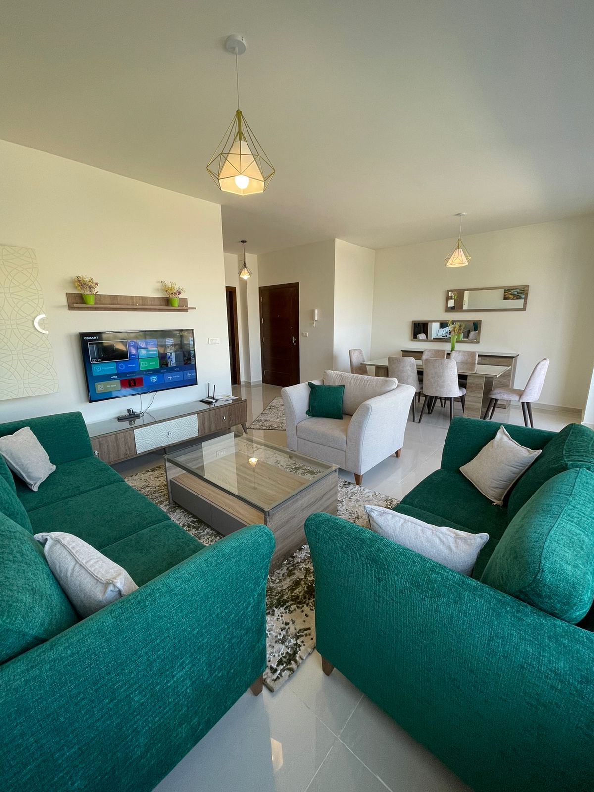 A modern living area is presented with comfortable green sofas arranged around a glass coffee table. A smart TV is mounted on the wall across from the seating. A dining table for four is visible in the background, illuminated by stylish pendant lights.
