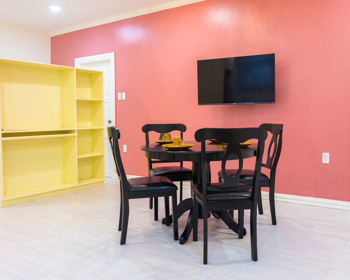 A dining area is presented with a round black table set for four, complemented by matching chairs. A wall-mounted television is visible, and a yellow bookshelf stands against the adjacent wall, enhancing the space's modern design. The room features a warm red accent wall.