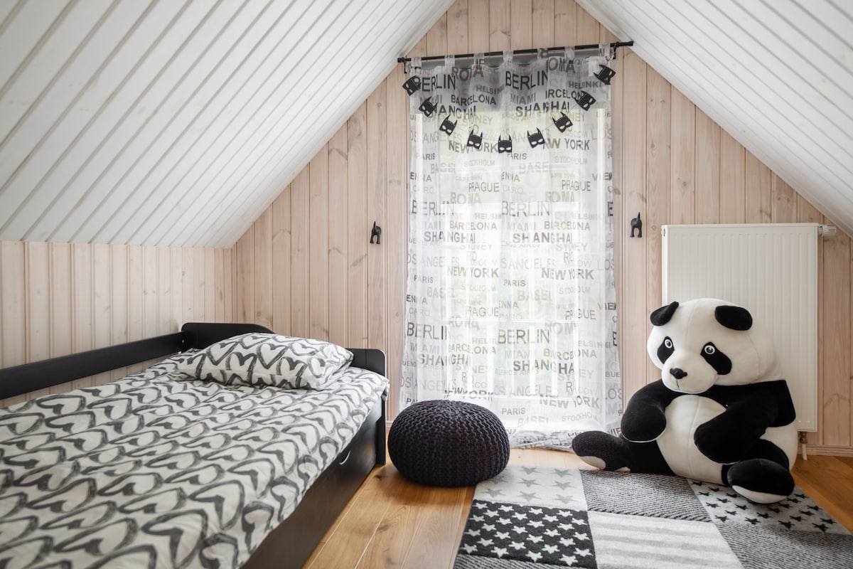 An inviting bedroom features a minimalist design with a simple bed positioned against light wooden walls. A large panda-shaped plush toy sits on a patterned rug beside a textured round pouf. Natural light filters through a curtain adorned with city names.