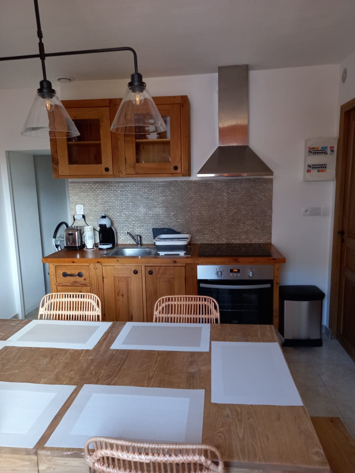 A warm kitchen area features wooden cabinetry and modern appliances. A table with four rattan chairs occupies the center, topped with placemats. A range hood is visible above the stove, and a coffee maker sits beside the sink, contributing to a functional cooking space.