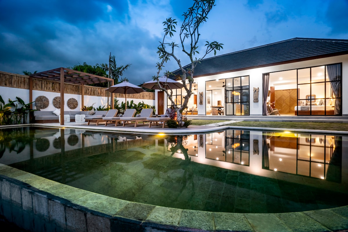 The exterior of the villa is visible at dusk, showcasing a private infinity pool reflecting the surrounding landscape. Sun loungers line the poolside, and a seating area under a shaded pergola is present. Lush tropical plants surround the area, enhancing the serene ambiance.