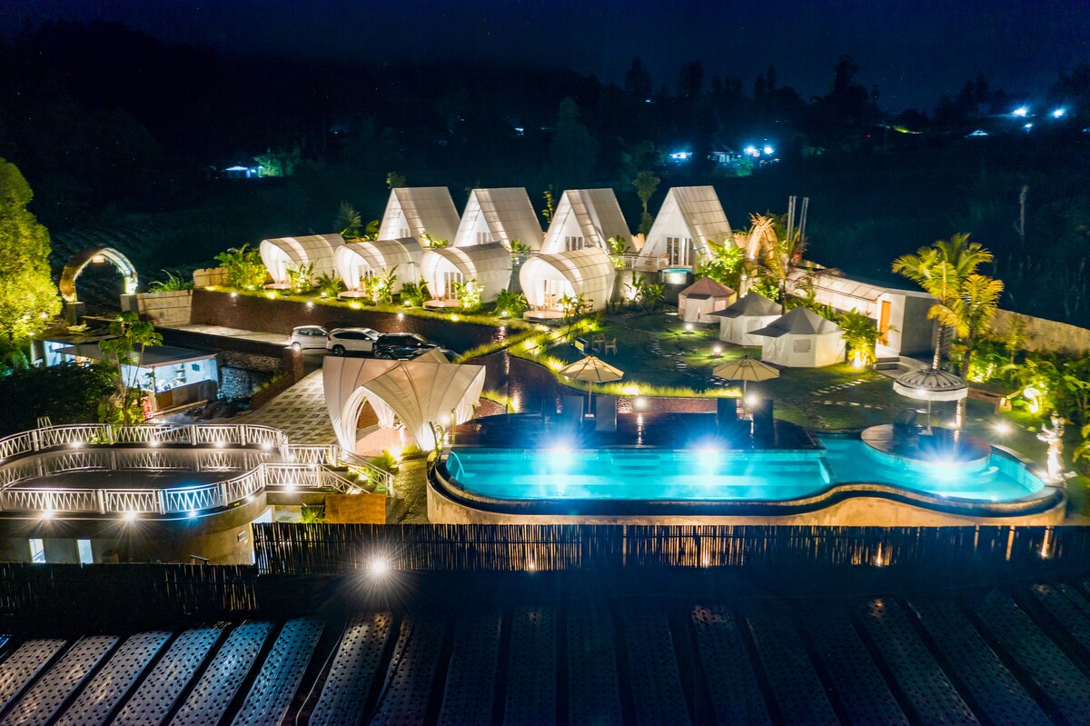 An aerial view showcases a unique glamping site with multiple tents arranged in a cluster. The area is illuminated at night, highlighting a swimming pool and landscaped gardens. Surrounding structures and pathways are visible, creating a serene environment in a natural setting.