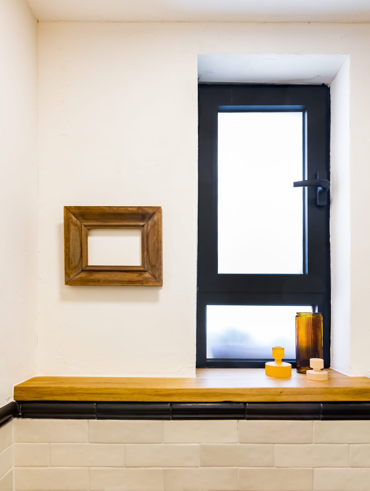 A minimalist corner of the bathroom features a wooden shelf placed under a narrow window. A wooden frame and two decorative glass vases are positioned on the shelf, contributing to a serene environment.