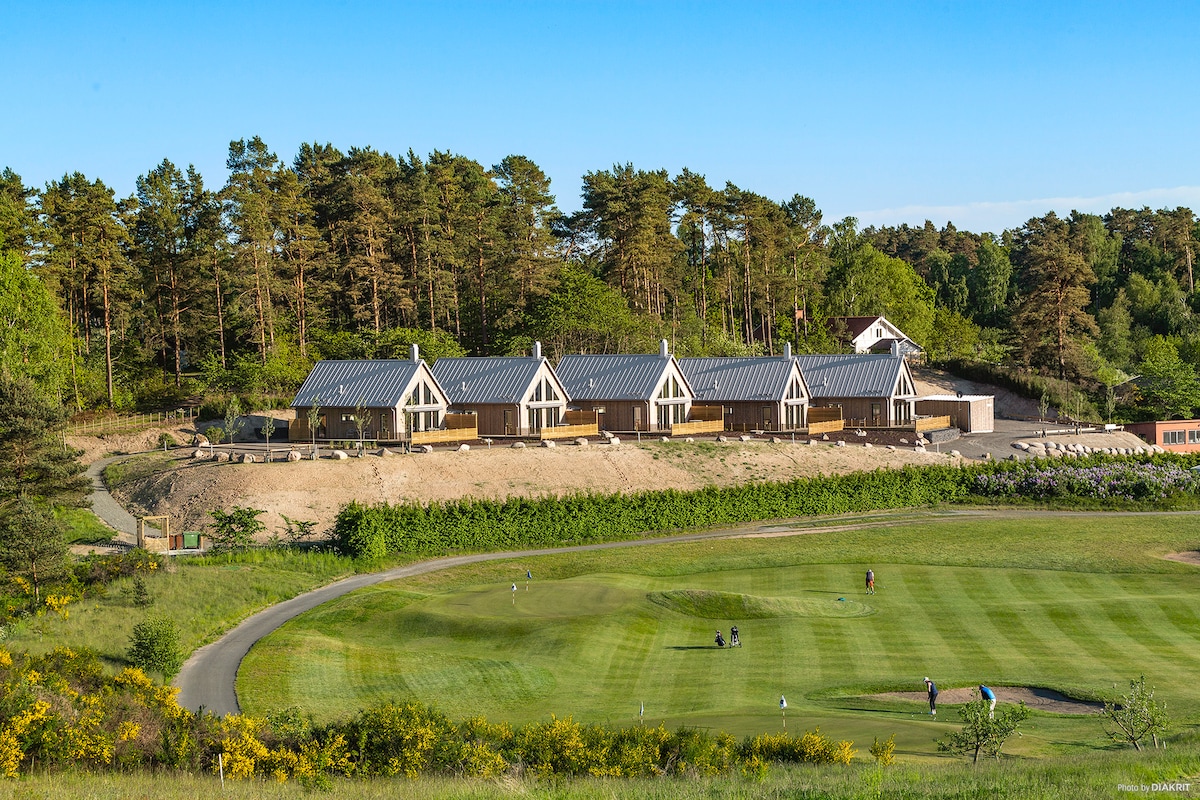 A row of modern villa units is positioned among trees, each featuring a sloped roof and large windows. An expansive green golf course is visible in the foreground, with people enjoying a game. Natural surroundings contribute to the serene environment.
